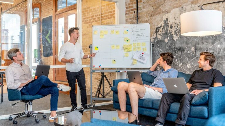 three men sitting while using laptops and watching man beside whiteboard